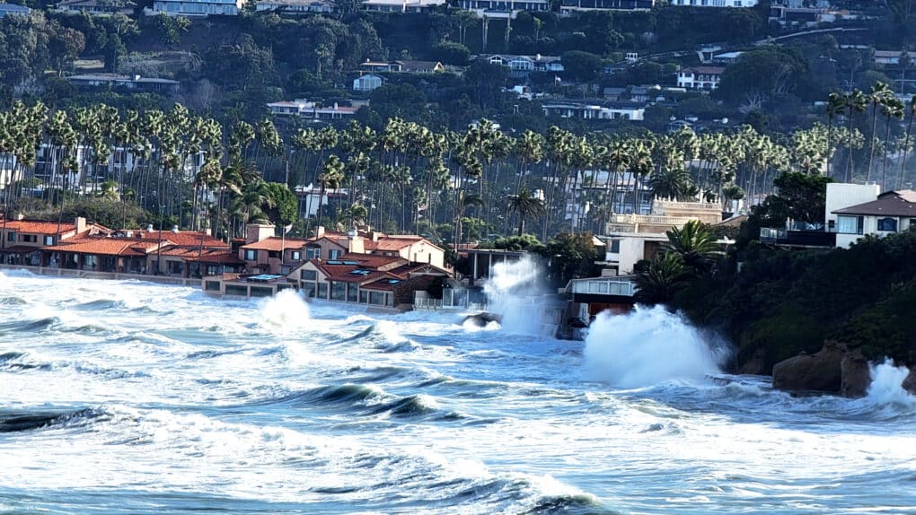 Waves are crashing against a series of homes with palm trees in the background, sending up water spray and waves. 