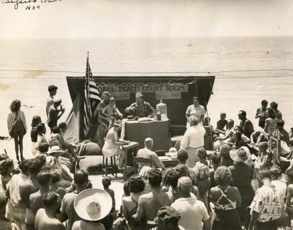 A large group of people are holding a mock court on the beach, complete with a judge, a large American flag, and a stenographer.