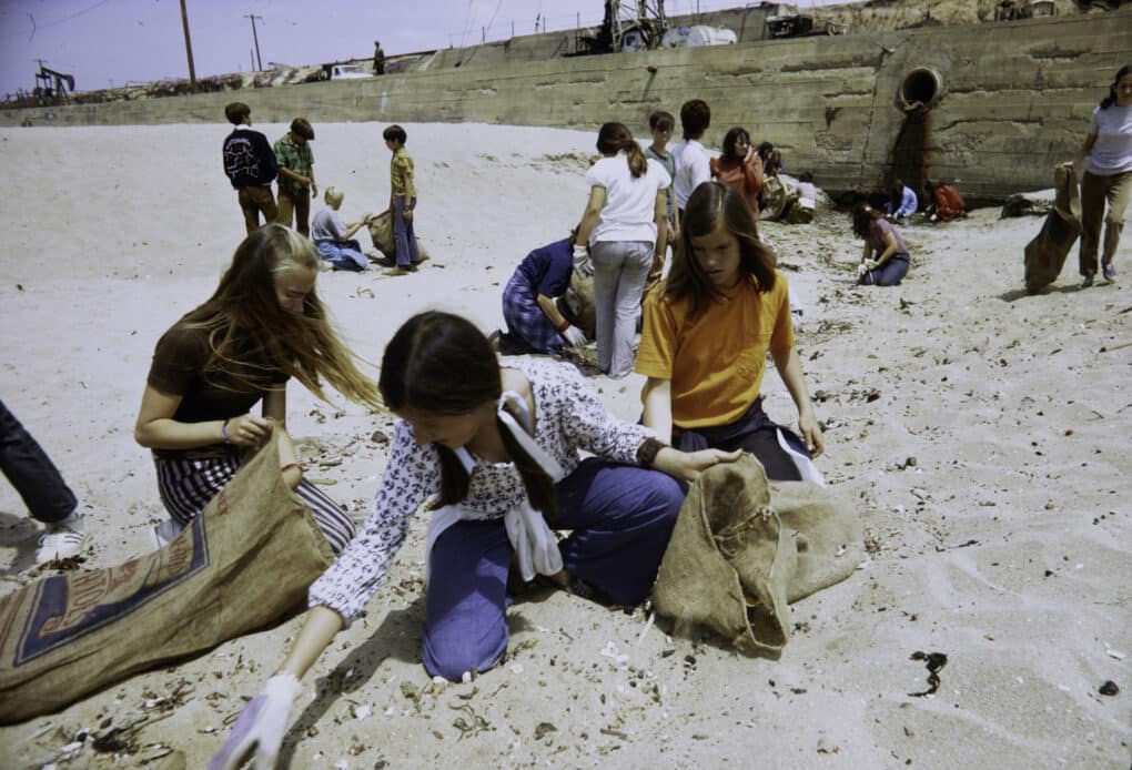 Middle-grade students are using burlap bags and wearing gloves to pick up a great deal of litter. Most are sitting or kneeling. 
