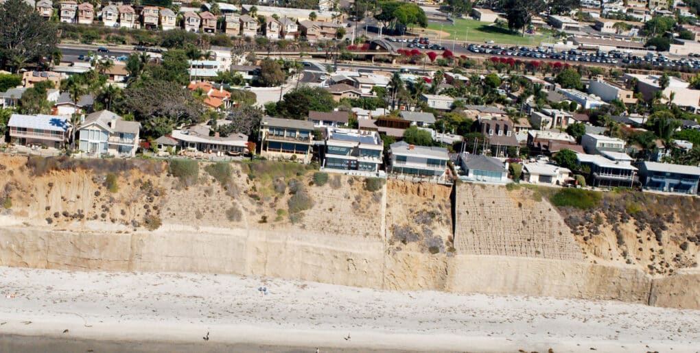 Aerial view of homes along a high sand bluff, with the ocean below. Sand bluff ends in sand-colored wall. 