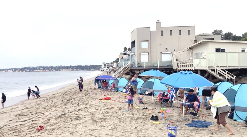 An all-ages group of people enjoying a cloudy day at the beach, with blue umbrellas and wind shelter tents on the sand.