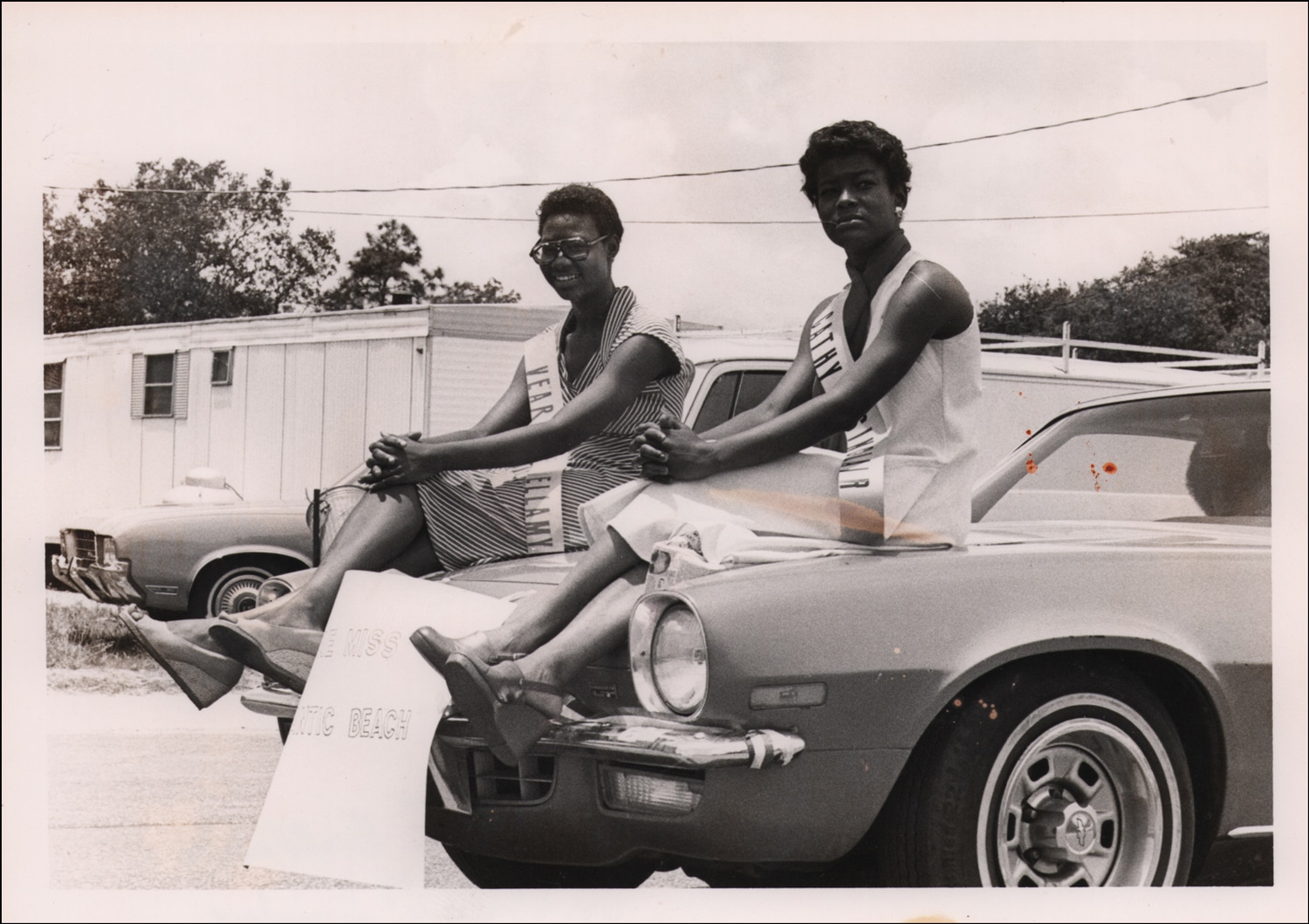 Click to Enlarge Black-and-white photograph of two young Black women in sundresses and sunglasses, wearing sashes, each sitting on the hood of a car.