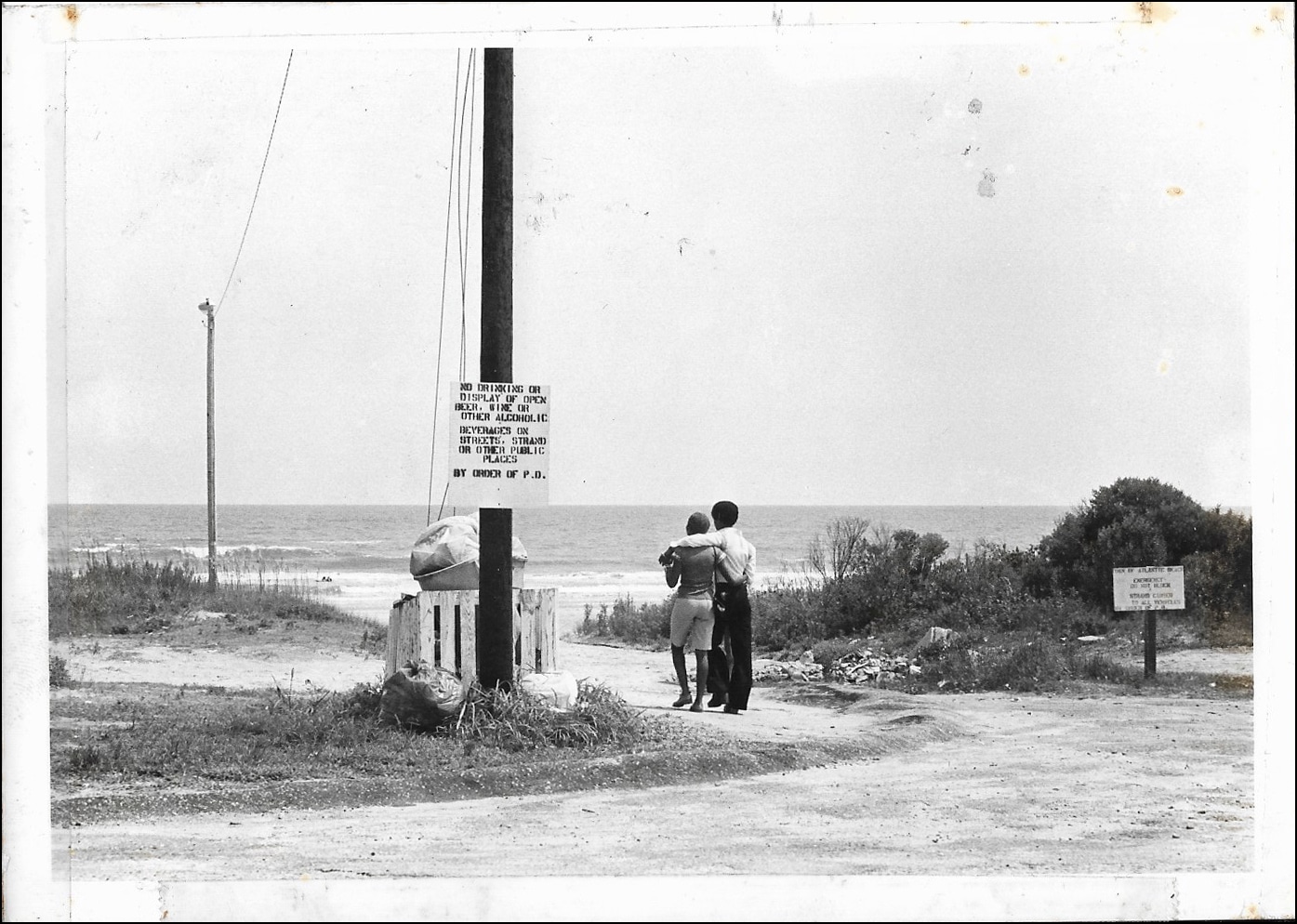 Click to Enlarge Black-and-white photo of young Black couple, seen from the back, walking on the beach.