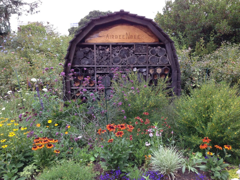 Color photograph of large wooden bee hotel surrounded by flowers. The wooden structure includes a wooden sign reading AIR BEE N BEE.