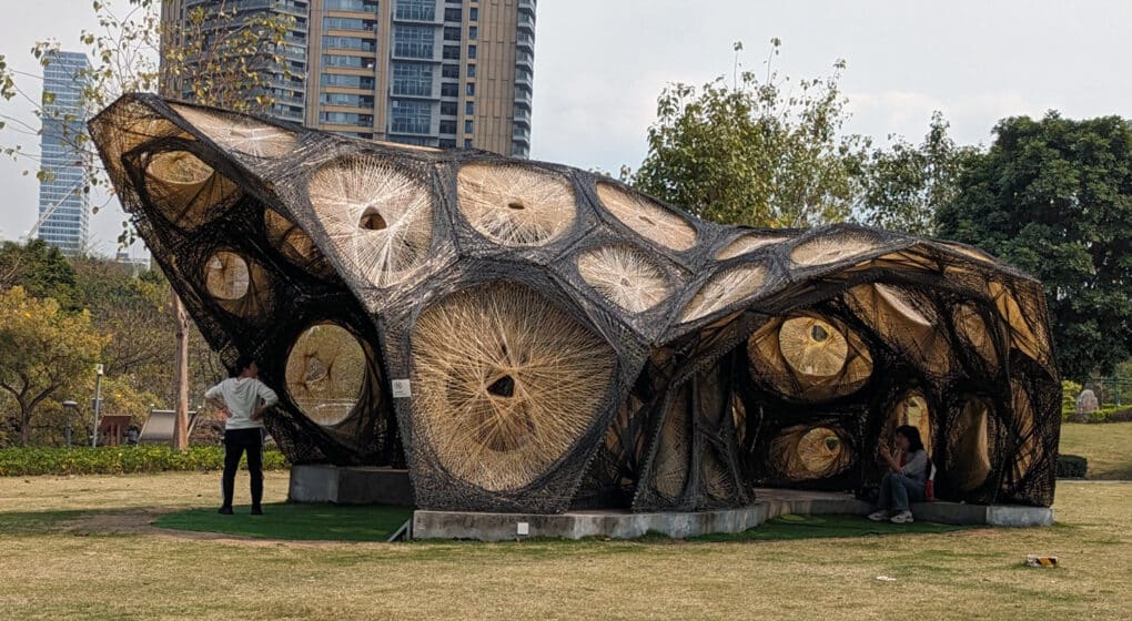 Color photograph of woven, biomorphic pavilion in a public park, with residential towers in the background.