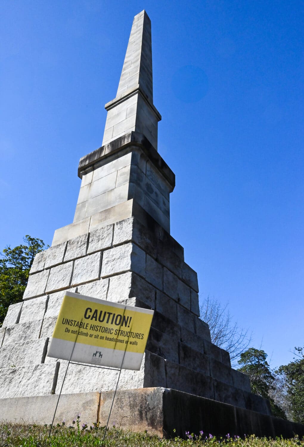Color photograph of a granite obelisk against blue sky, with a sign in front of it reading "CAUTION: Unstable historic structures. Do not climb or sit on headstones or walls."