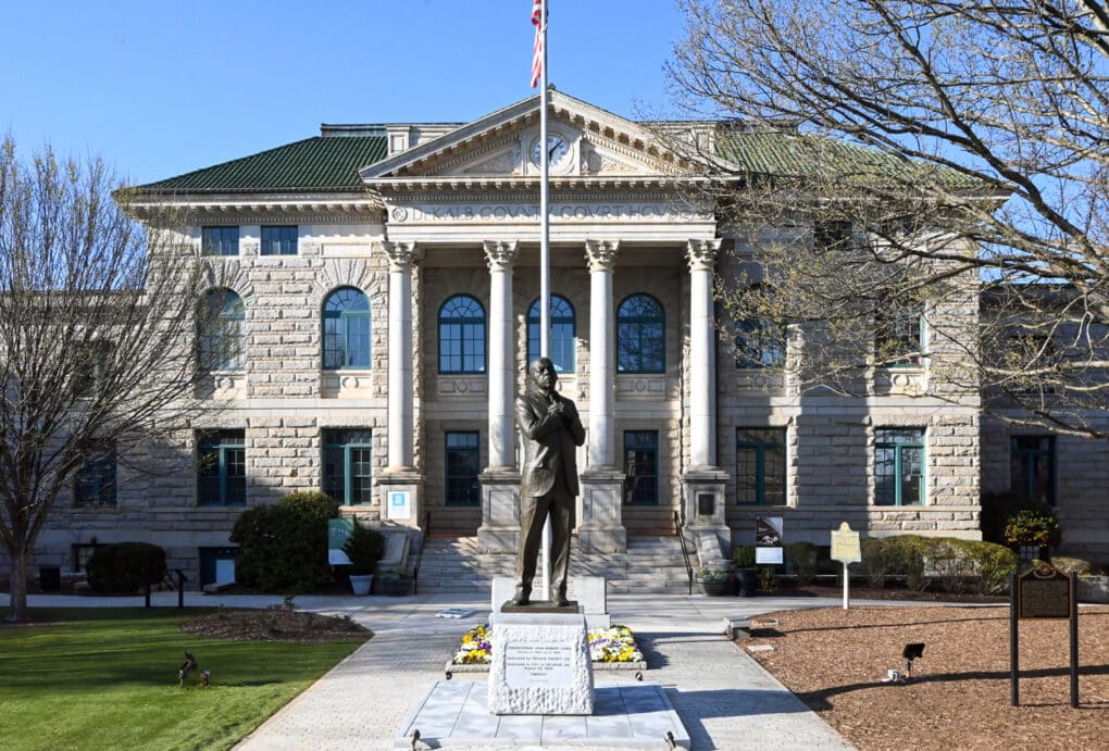 Color photograph of a neoclassical civic building, in front of which stand an American flag on a flagpole and a bronze statue of a middle-aged Black man wearing a business suit.