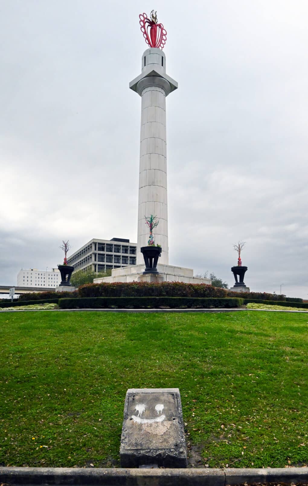 Color photograph of monumental obelisk topped with pink filigree heart, surrounded by green lawn. In the foreground is a concrete block on which has been spray-painted a smiley face.