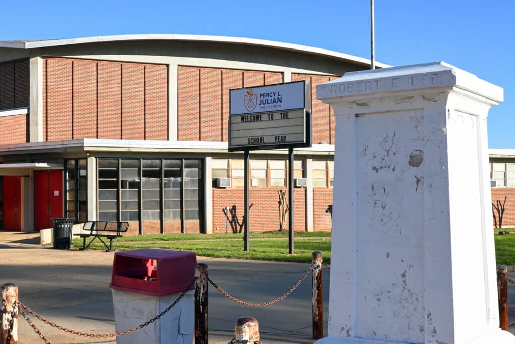 Color photograph of a brick school building, with a sign in front reading "Percy L. Julian High School." In the foreground is an empty plinth, covered in peeling white paint and inscribed "Robert E. Lee."