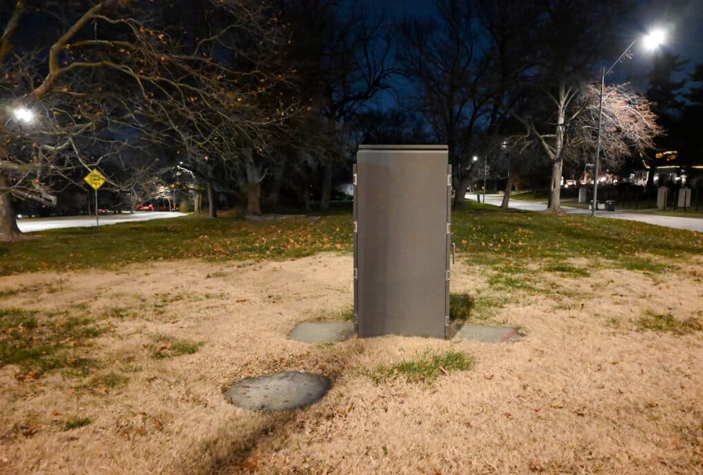 Color photograph of a tall metal electrical box in a small city park at night.