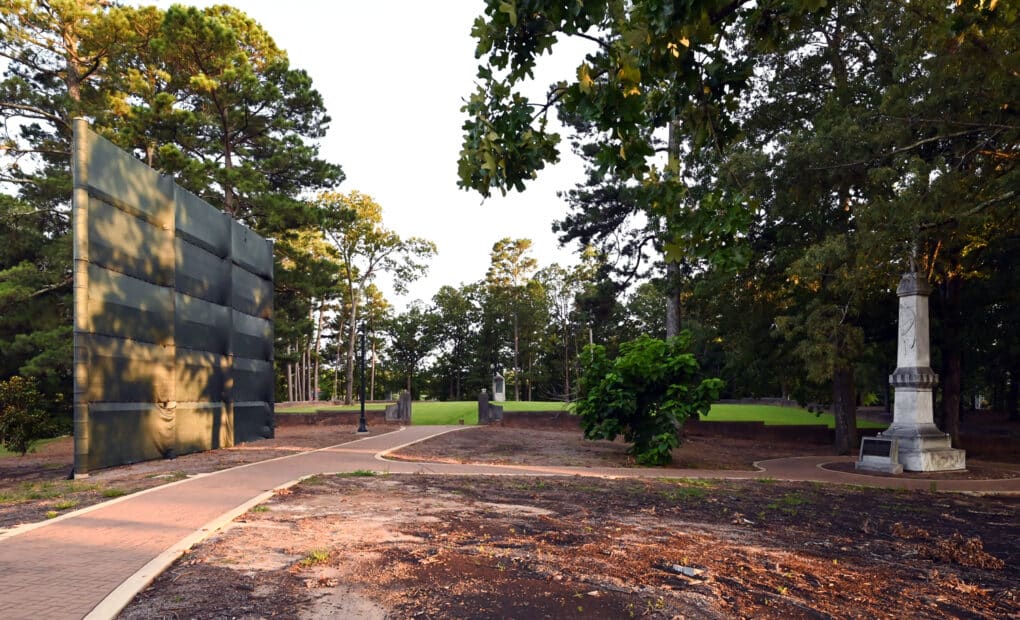 Color photograph of a leafy walkway on a college campus; to the right is a plinth topped by a statue; to the left is a tall screen made of environmental fabric.
