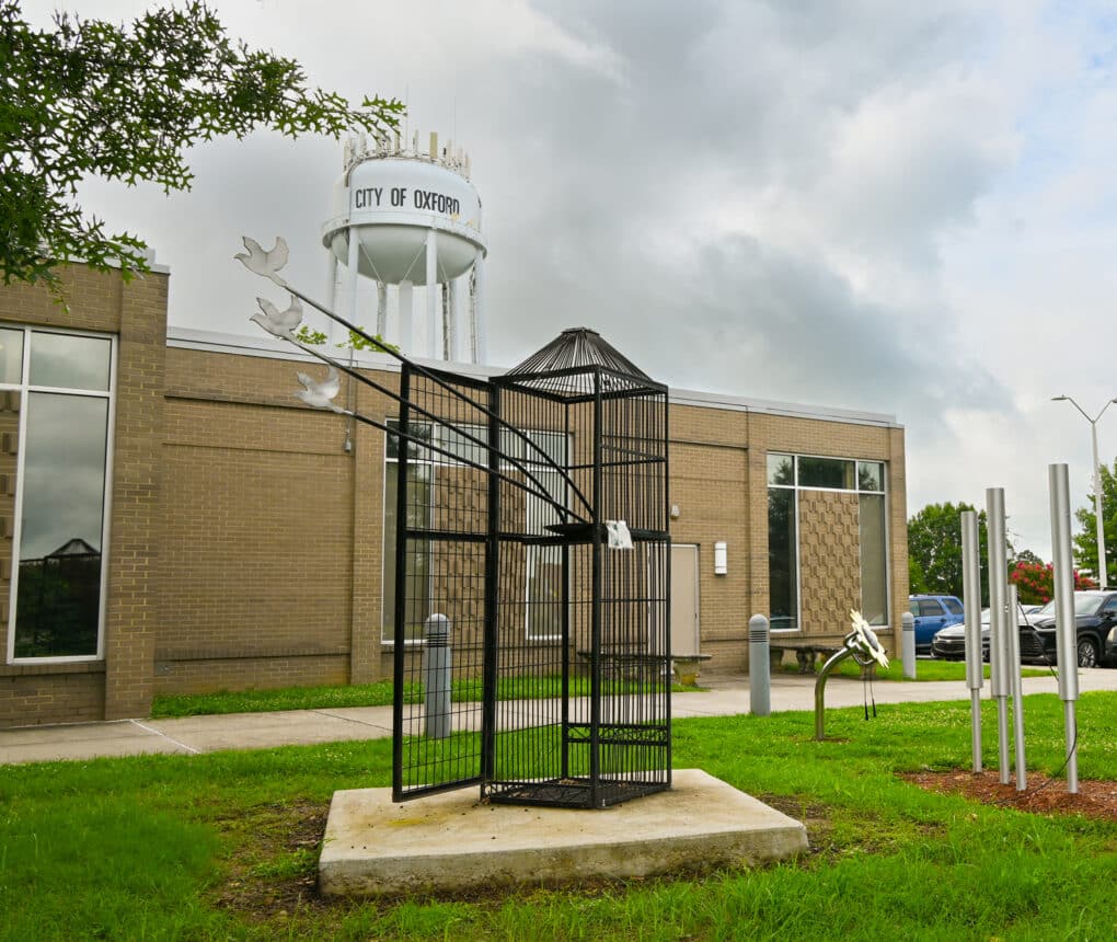 Color photograph of beige-brick municipal building. In the foreground is a sculpture depicting a giant black birdcage, with three metal doves emerging from it; in the background is a water tower with the words CITY OF OXFORD.