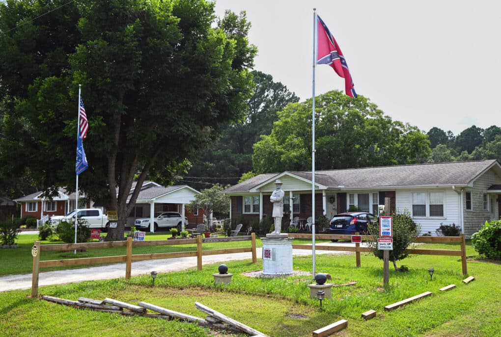 Color photograph of a small Civil War statue, situated on the lawn of a suburban ranch-style house. Beside it are American and Confederate flags on flagpoles.