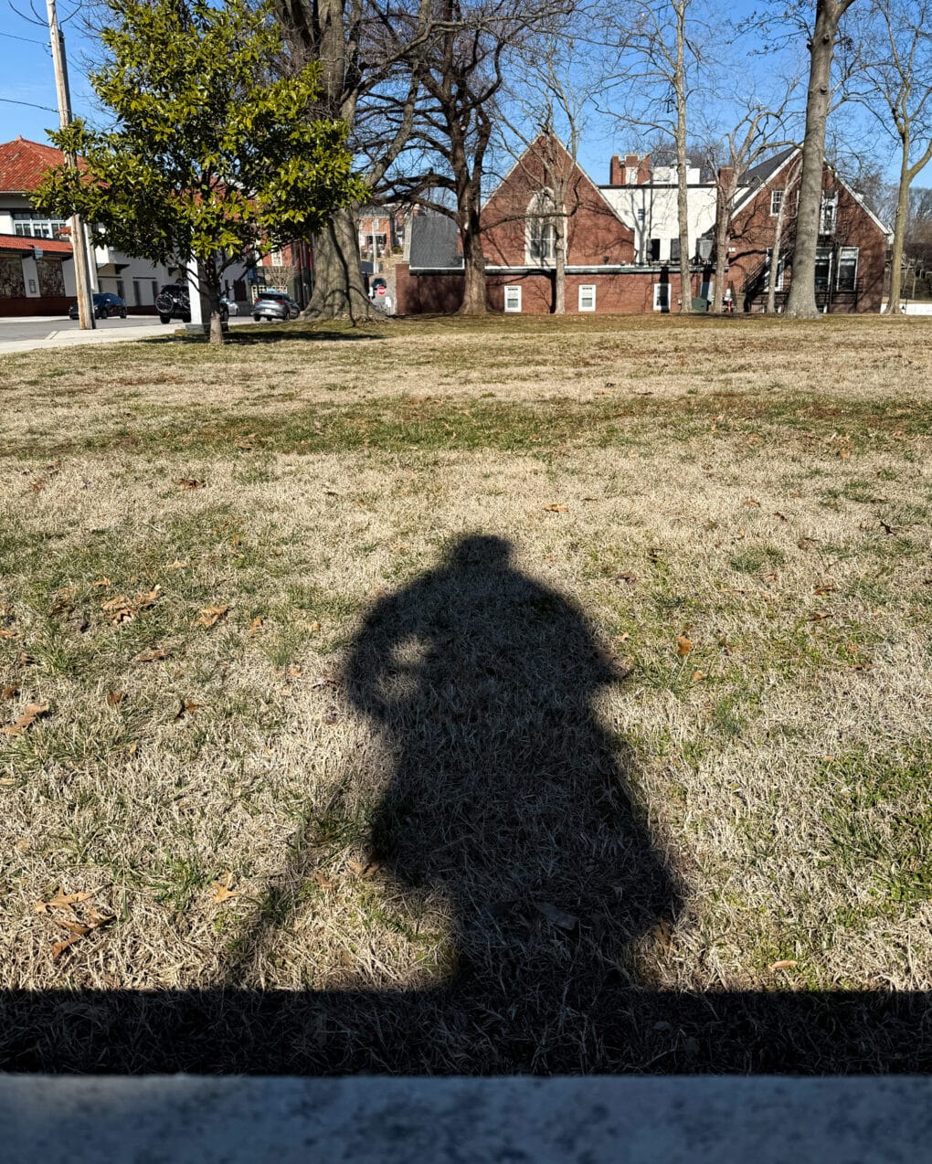 Color photograph of the shadow of a Civil War soldier statue, cast on the grass in a park.
