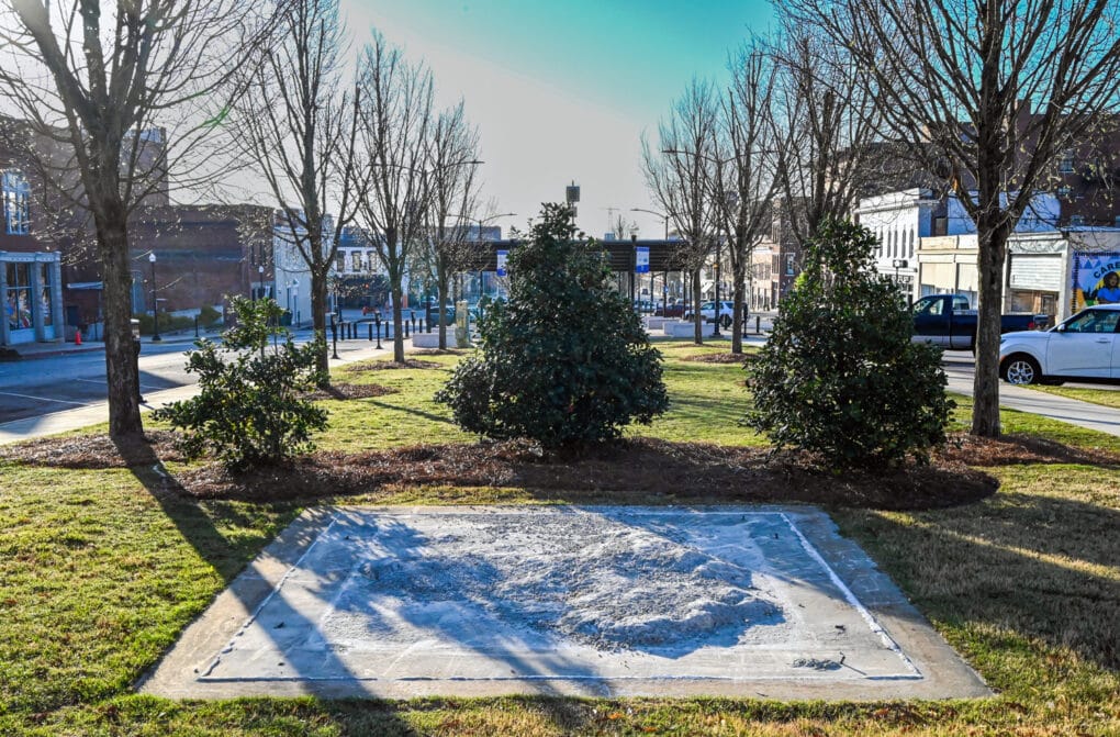 Color photograph of a cement square, rubble in its center, situated in a small park in the center of what appears to be an old-fashioned downtown.