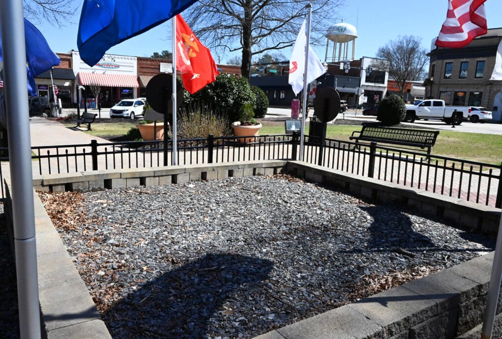 Color photograph of a cement-walled, gravel-paved enclosure, flanked by flags and situated in a small park in the center of what appears to be an old-fashioned downtown.
