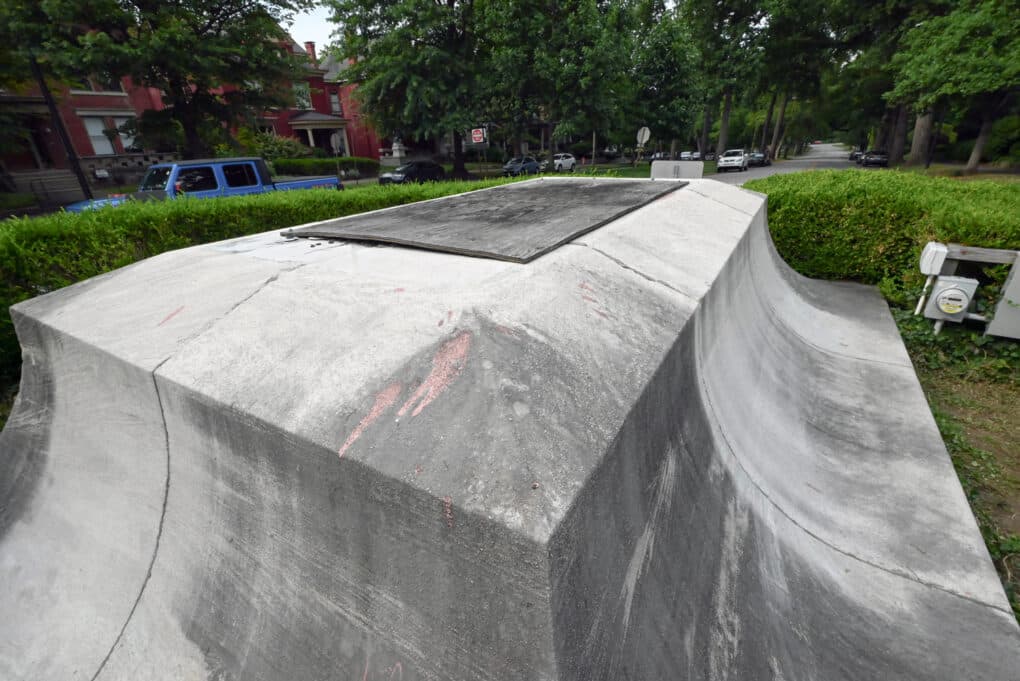 Color photograph of a cement statue base, topped with a weathered piece of plywood, in a leafy residential neighborhood.