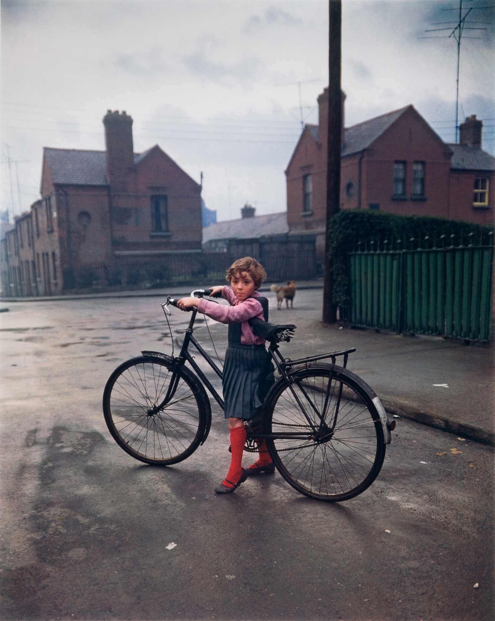 Color photograph of small girl straddling an adult-sized bicycle. She wears a pink shirt, blue jumper, and red socks; behind her are brown brick tenement houses and a cloudy sky.