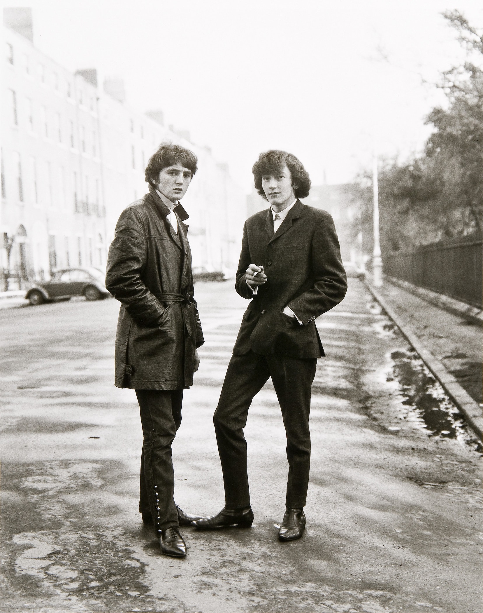Black-and-white photograph of two young white men, wearing skinny black pants, winkle-picker boots, and long jackets, standing on a city street.