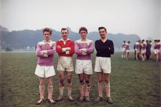 Color photograph of four young men standing in a row, arms folded, on a green grassy sports field. They wear white shorts, colorful jerseys, dirty ankle-length socks, and football shoes, and are smiling at the camera.