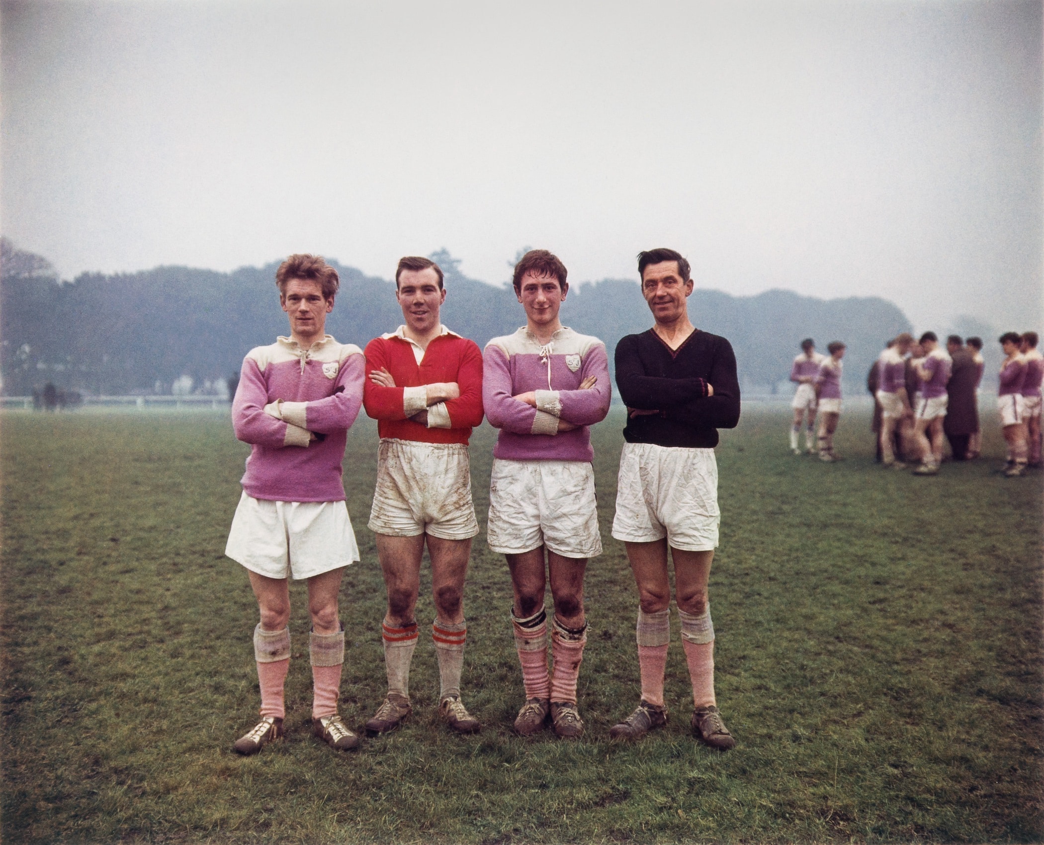 Color photograph of four young men standing in a row, arms folded, on a green grassy sports field. They wear white shorts, colorful jerseys, dirty ankle-length socks, and football shoes, and are smiling at the camera.