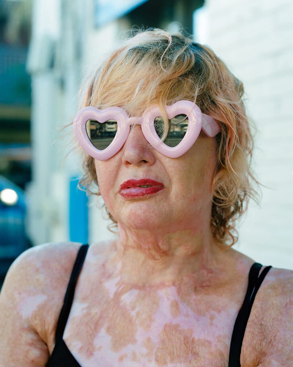 Color photograph of middle-aged white woman with blonde hair, large pink heart-shaped sunglasses, black tank top, and red lipstick.