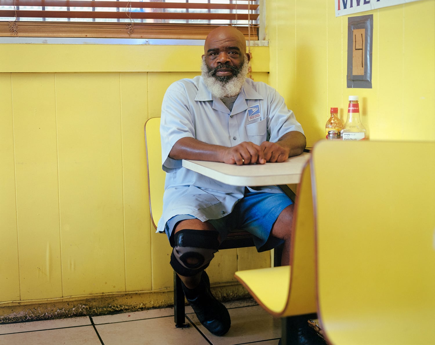 Click to Enlarge Color photograph of middle-aged Black man in postman's uniform, seated at a restaurant table in yellow-painted interior.