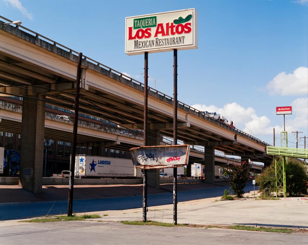 Color photograph of a sign reading TAQUERIA LOS ALTOS MEXICAN RESTAURANT beside an elevated freeway.