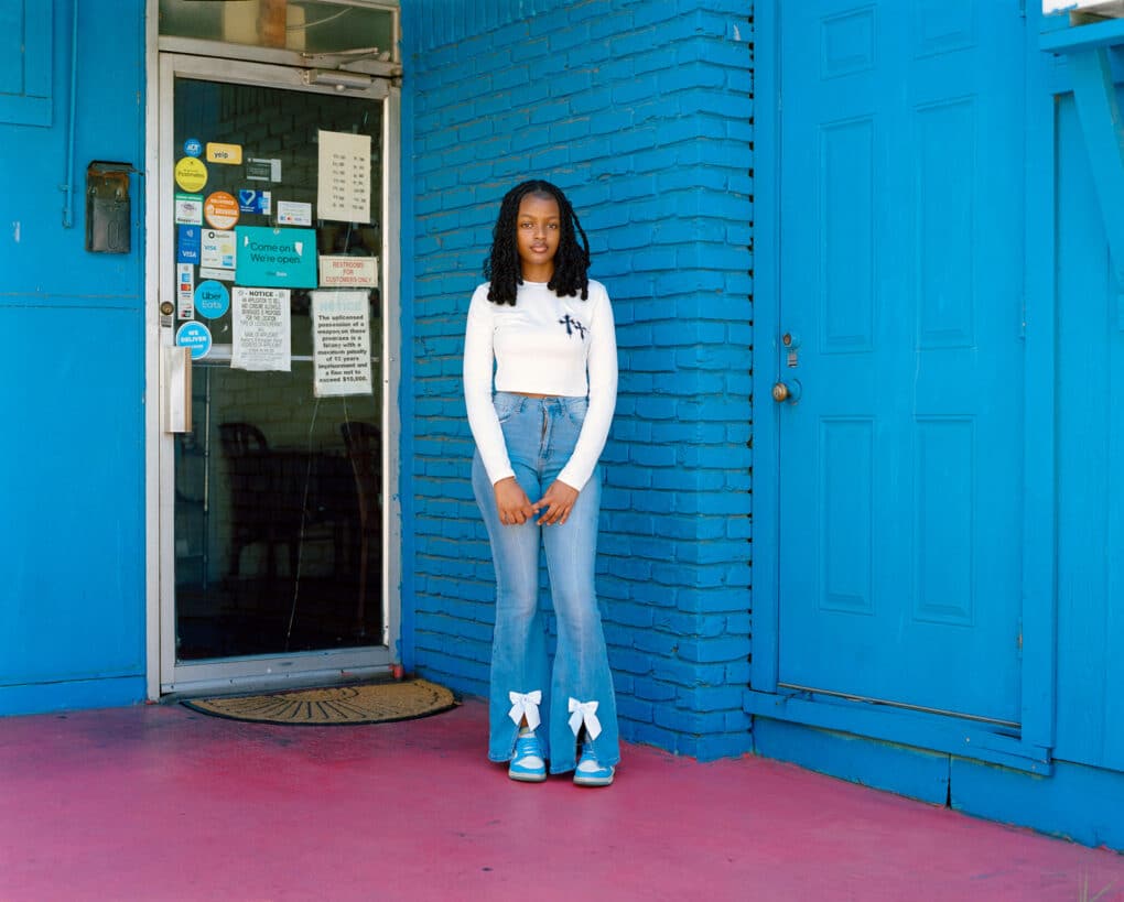 Color photograph of Black teenage girl in jeans and white shirt, standing in front of blue-painted wall.