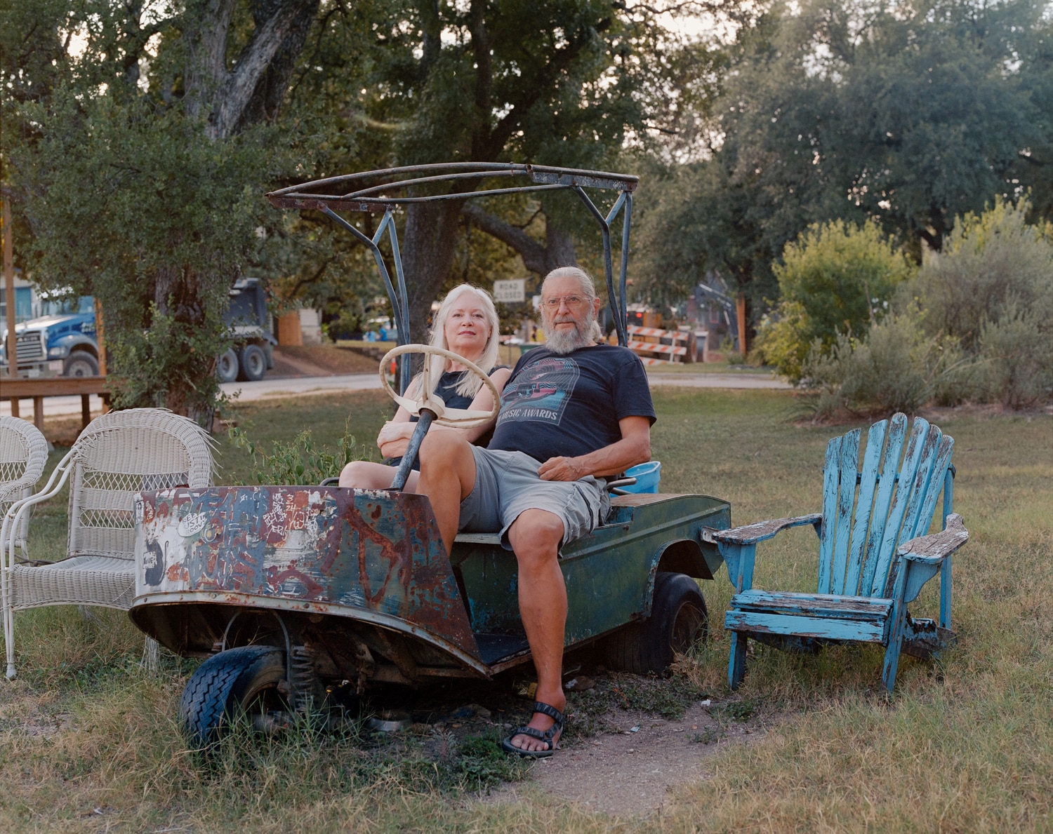 Click to Enlarge Color photograph of a middle-aged white couple on a beat-up golf cart, with trees and grass in the background.