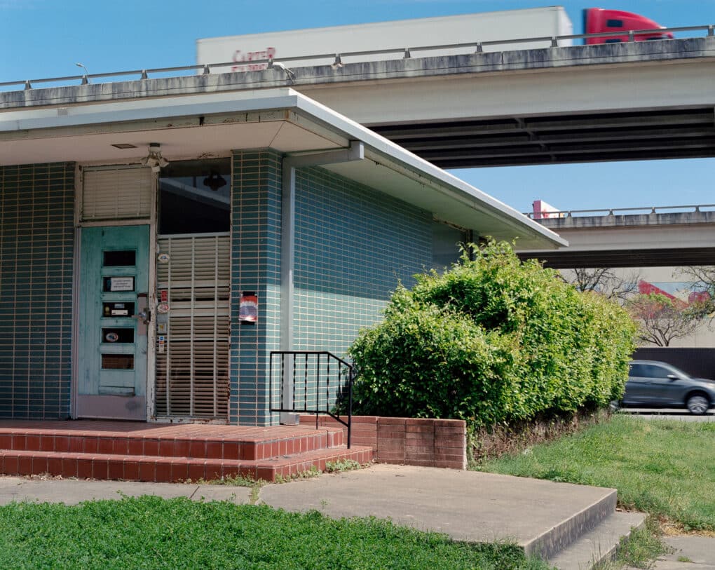 Color photograph of a one-story building sheathed in green tiles beside an elevated highway.