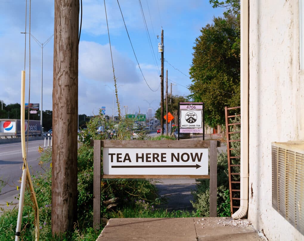 Color photograph of a sign along a roadway, reading TEA HERE NOW.
