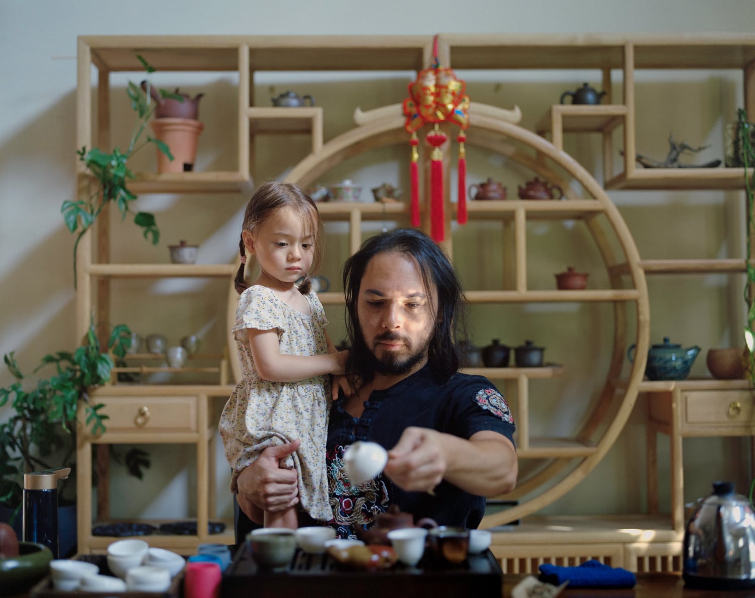 Click to Enlarge Color photograph of young Asian man and small Asian girl in an interior; the man is pouring a cup of tea.