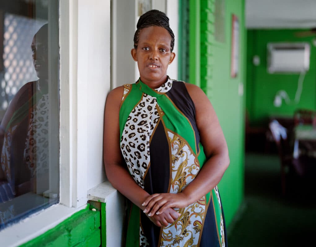 Color photograph of Black woman wearing green and black dress, standing outside green-painted restaurant.