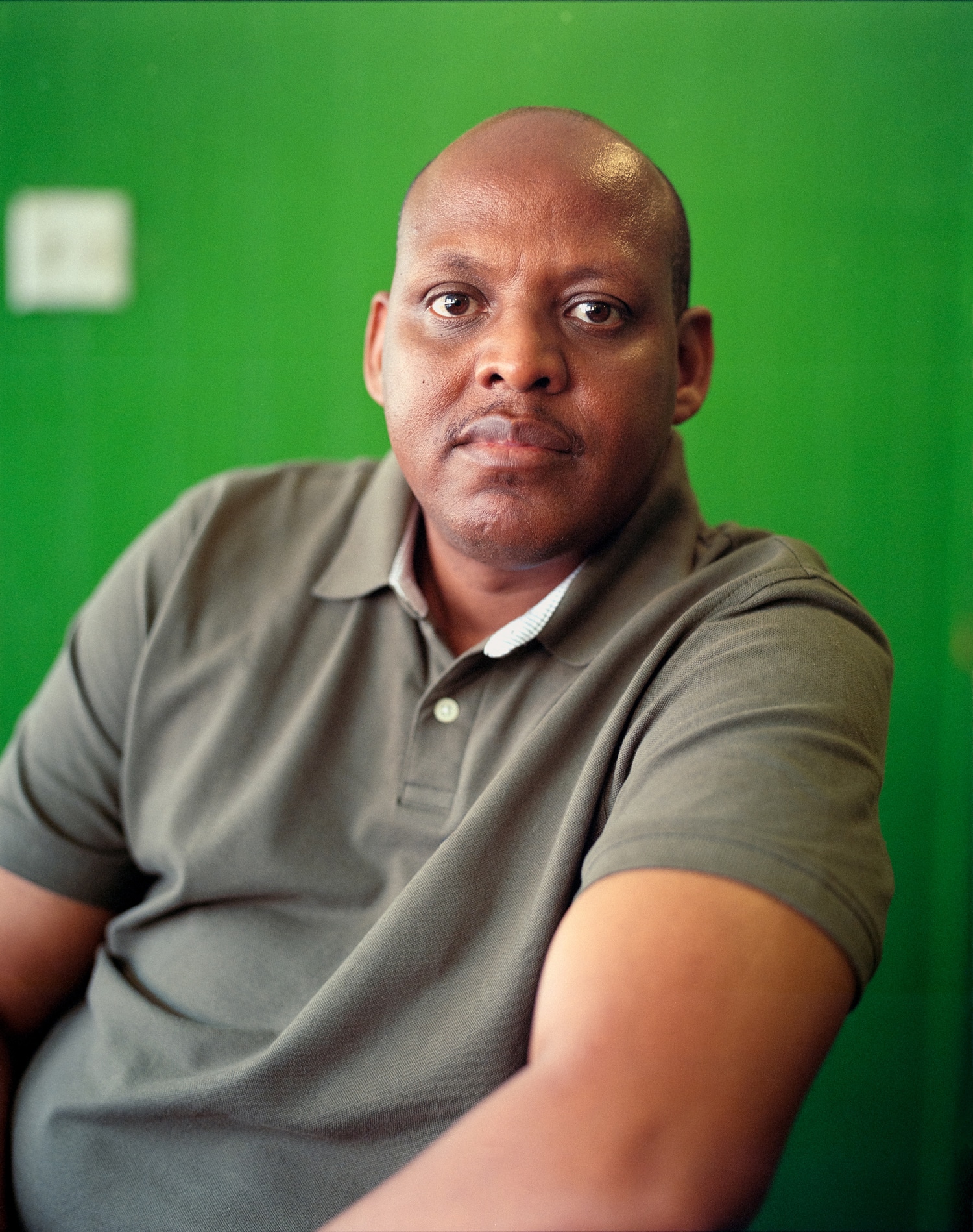 Click to Enlarge Color portrait of balding Black man in green polo shirt, looking directly at the camera.