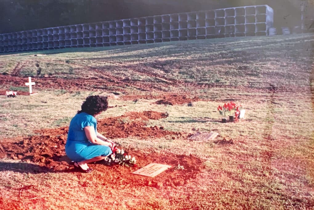 Color photograph of middle-aged Chamorro woman crouching down beside a headstone.