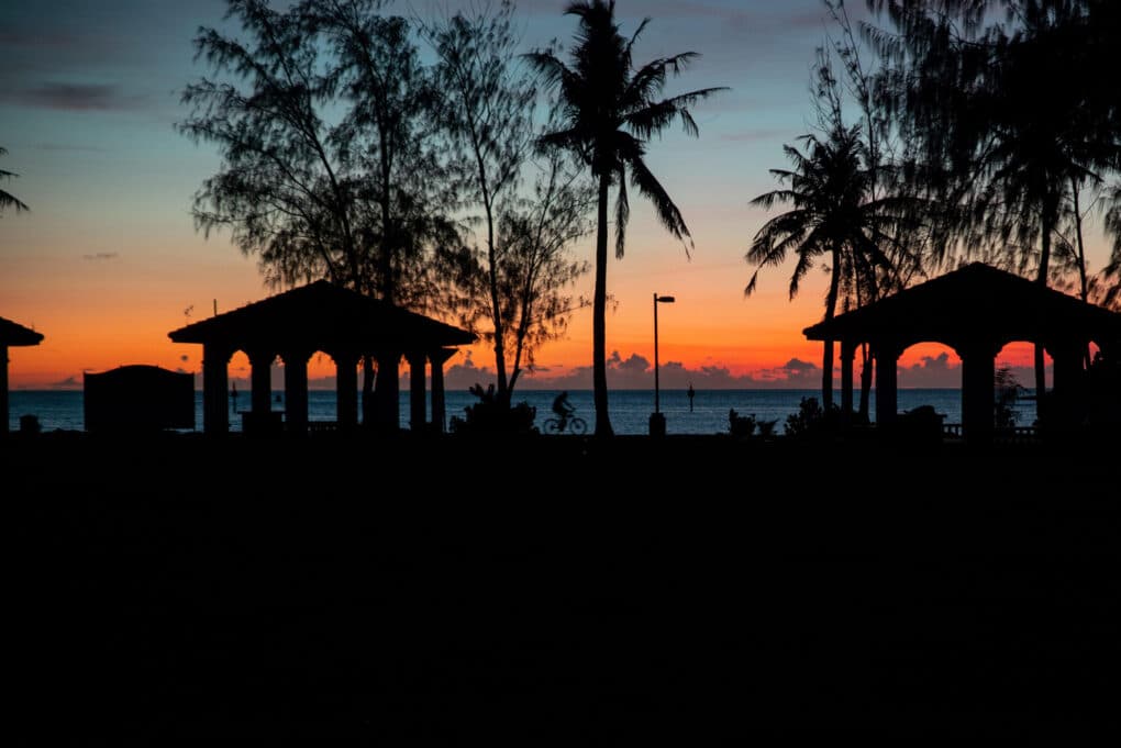 Color photograph of red sunset over water, with silhouetted trees and picnic pavilions in the foreground.