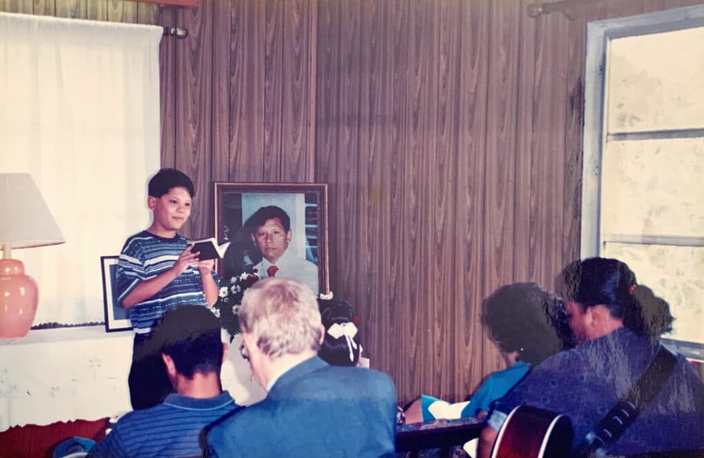 Color photograph of a boy in a striped shirt standing in a domestic interior, holding a book in his hands and looking out at seated guests; behind him is a framed photograph of a man in a suit.