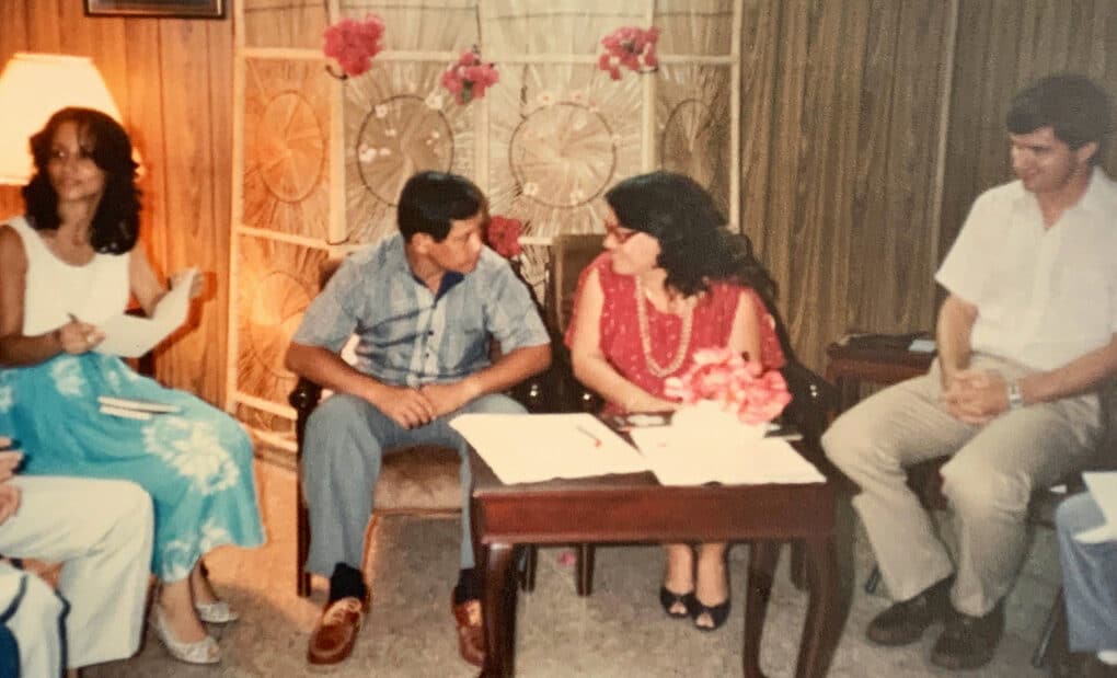 Color photograph of a Chamorro couple seated at a small table in a domestic space, with guests seated around them.