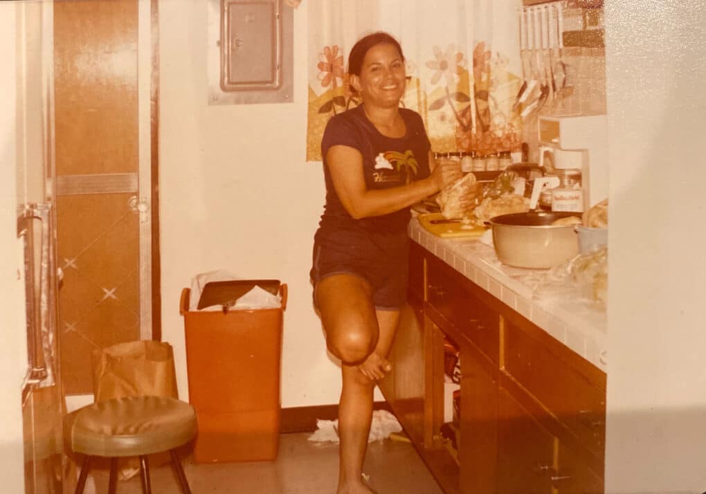 Color photograph of young woman in shorts and t-shirt, barefoot, cooking in her kitchen.