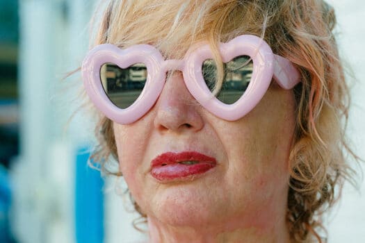 Color photograph of middle-aged white woman with blonde hair, large pink heart-shaped sunglasses, and red lipstick.