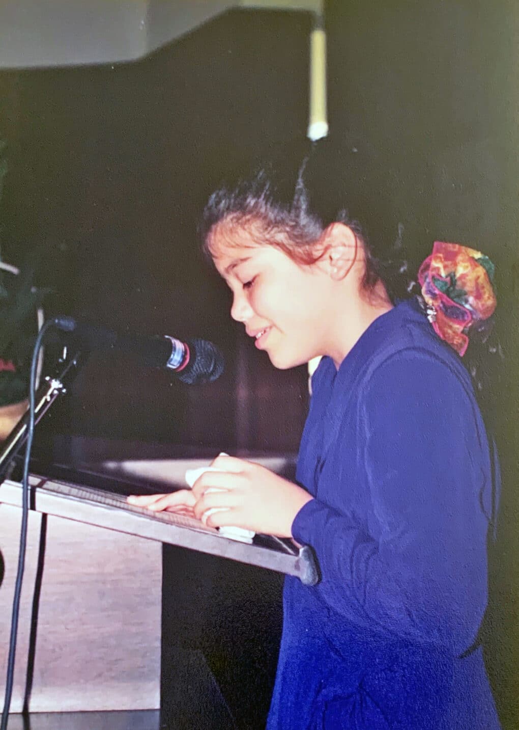 Color photograph of a small girl with dark hair, wearing a dark blue dress, reading at a podium.