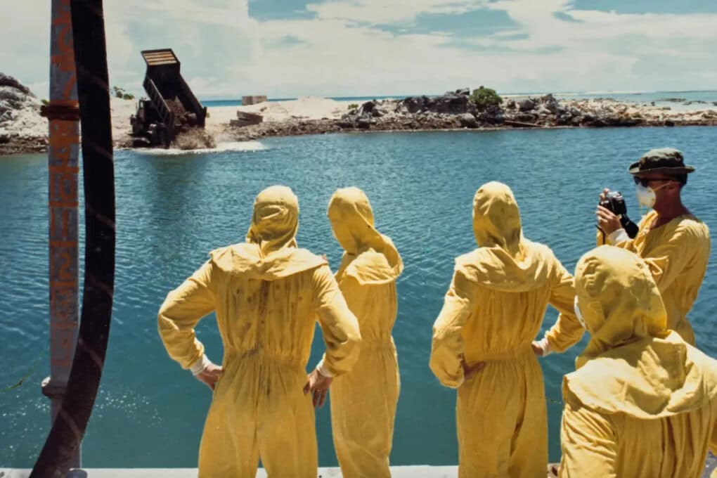 Color photograph of five men in yellow hazmat suits, watching a dump truck pitch material into the water.