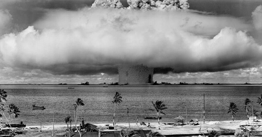 Black-and-white photograph of a huge mushroom cloud, with palm trees along a beach in the foreground.