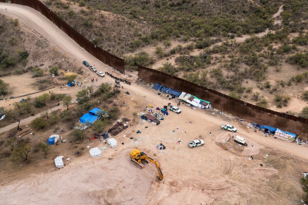 Aerial photo of border wall with small gap. There are several vehicles, some tents, a line of people, barely visible from distance. 