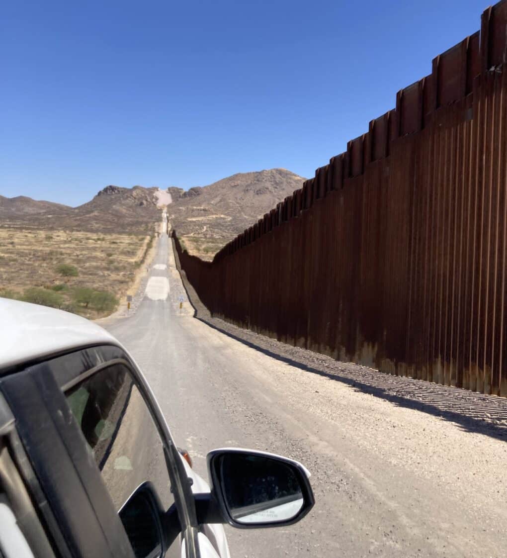 Photo taken from white car with bollards of border wall, rust-colored to right, alongside gravel road, approaching an incline.