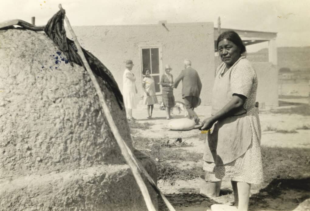 A Native woman is standing in front of a large mound of adobe, holding a loaf of bread on a flat board. 