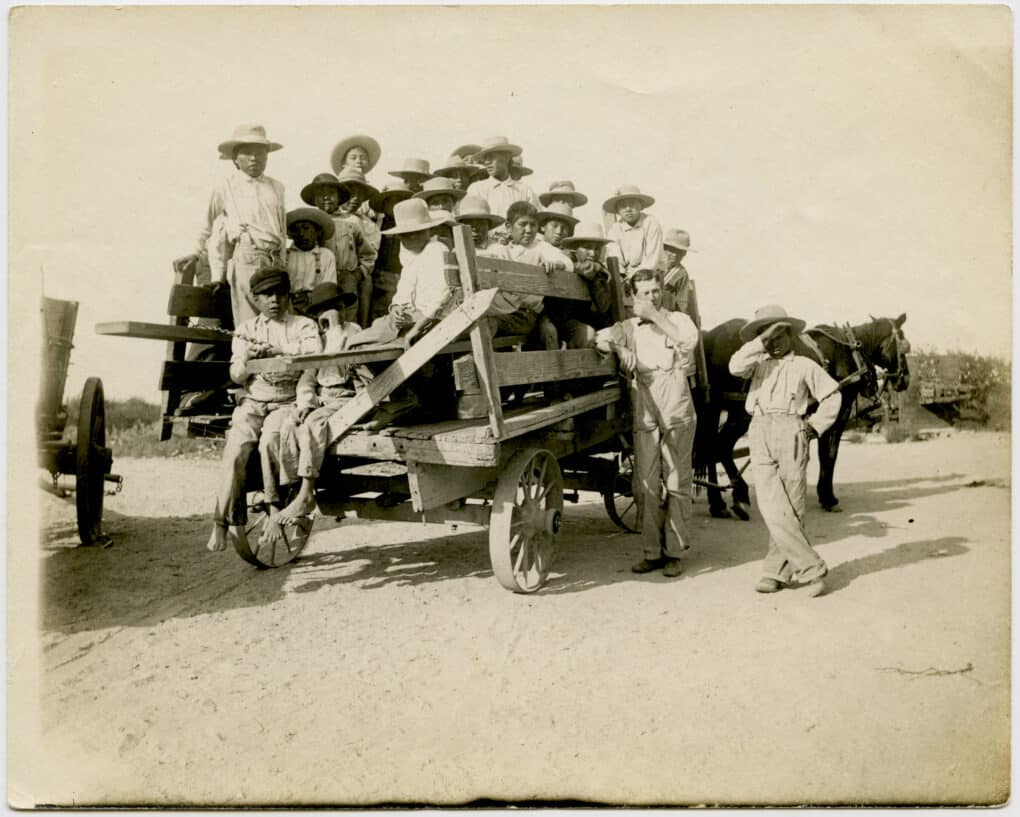 Sepia-tone photo of many young boys wearing straw hats piled in the back of a ramshackle truck on a dirt road, and an adult man giving a thumbs up signal. 