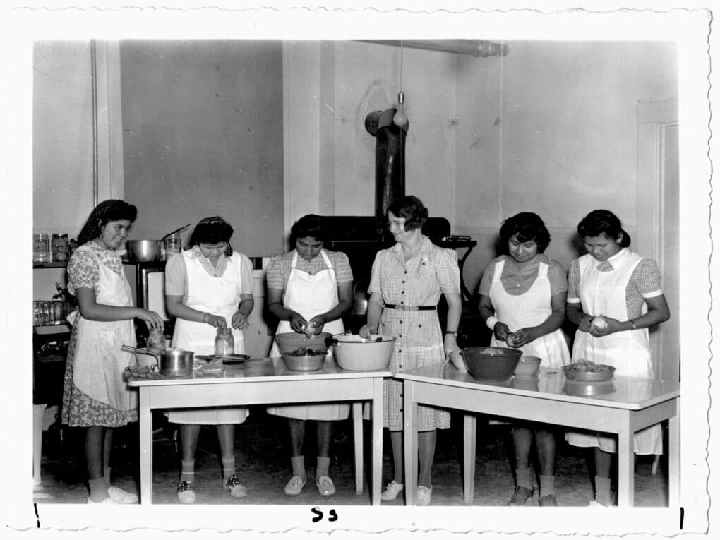 Black and white photo showing five teenage girls apparently cooking with aprons on, and a teacher in the middle, supervising. 