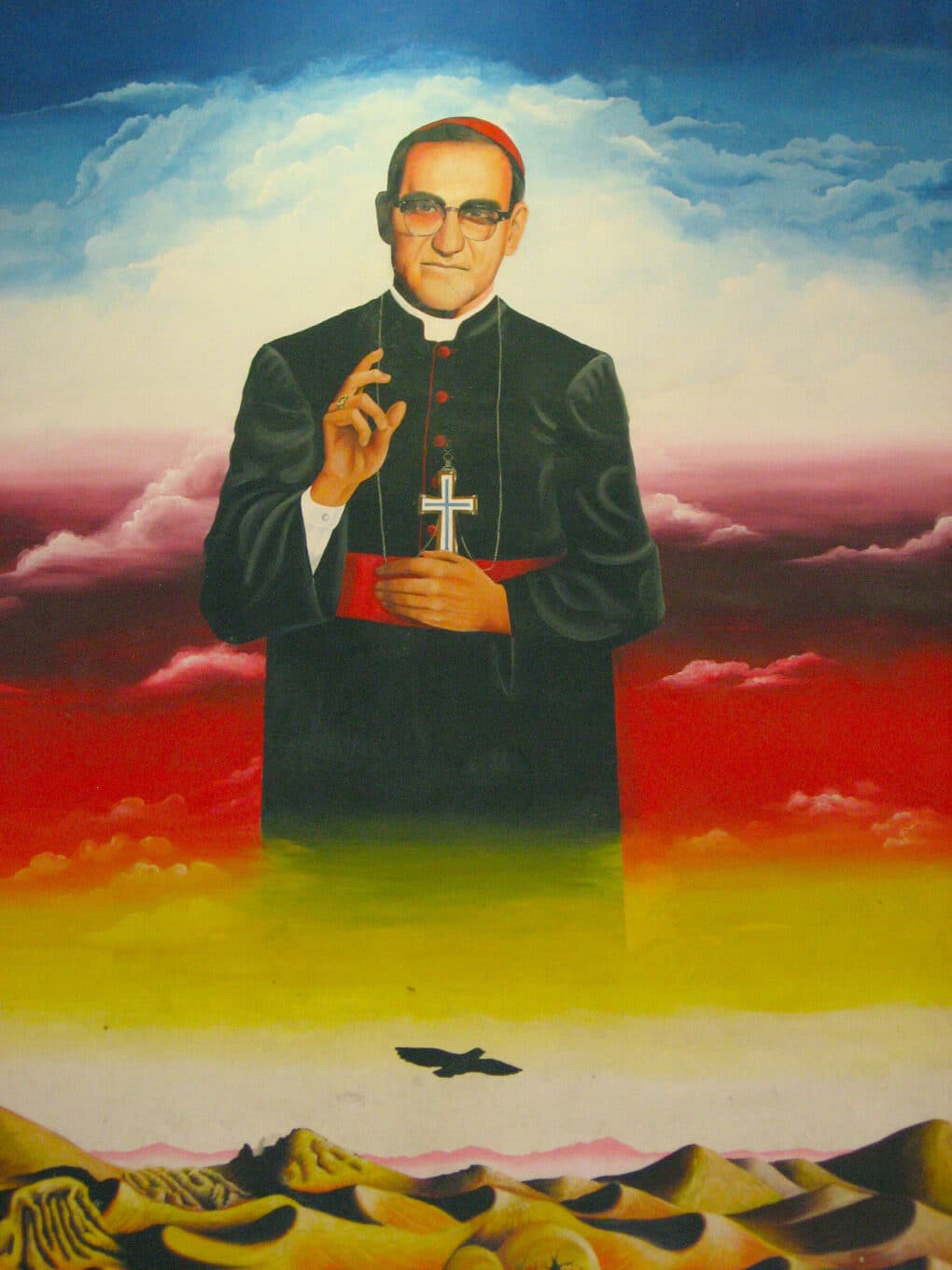 Brightly colored vertical painting, possibly a mural, of priest Óscar Romero in vestments, with backdrop of desert. 